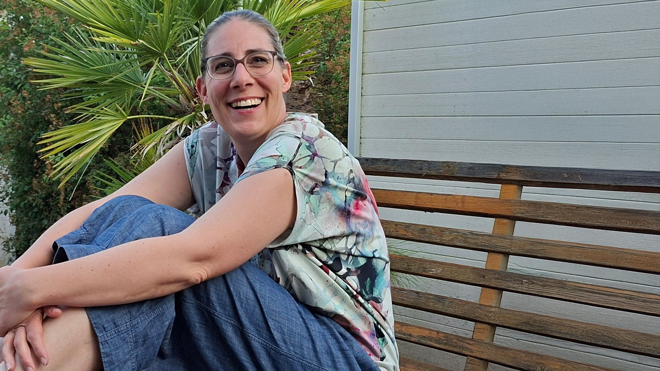 Eine fröhliche Frau sitzt auf Gartenbank und schaut in die Kamera | © Familie Wirz