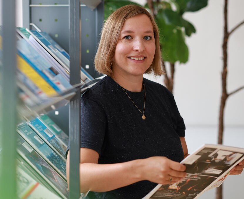 Snežana Milojević steht an einem Regal, lacht in die Kamera und hält eine Broschüre in der Hand | © ask! – Beratungsdienste für Ausbildung und Beruf