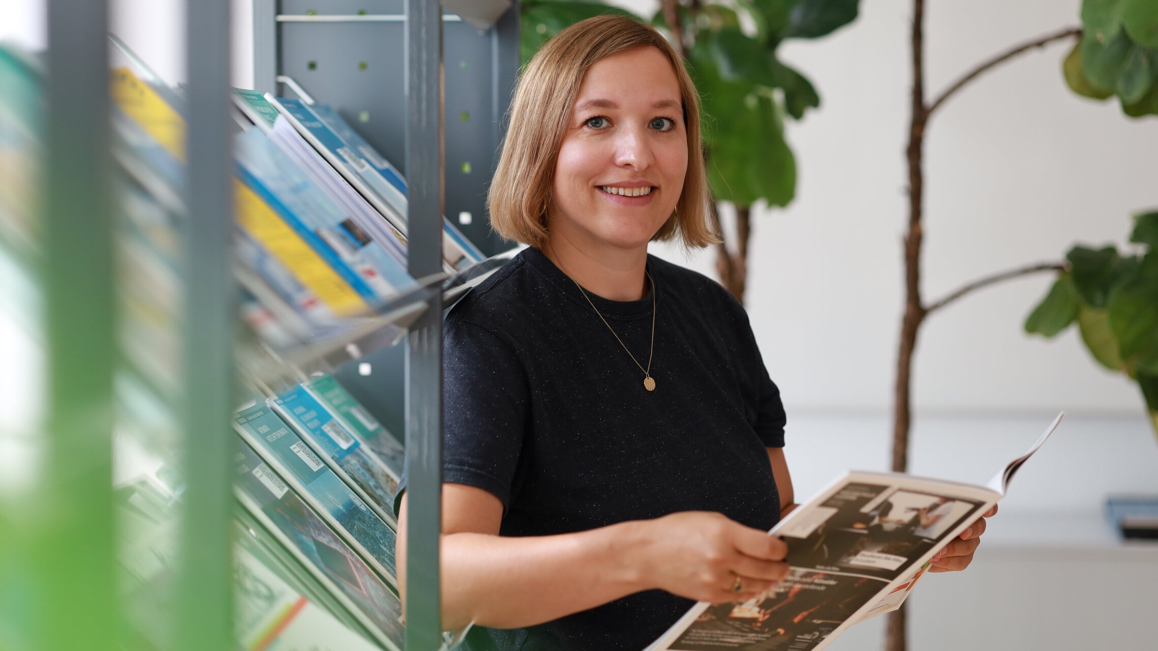 Snežana Milojević steht an einem Regal, lacht in die Kamera und hält eine Broschüre in der Hand | © ask! – Beratungsdienste für Ausbildung und Beruf