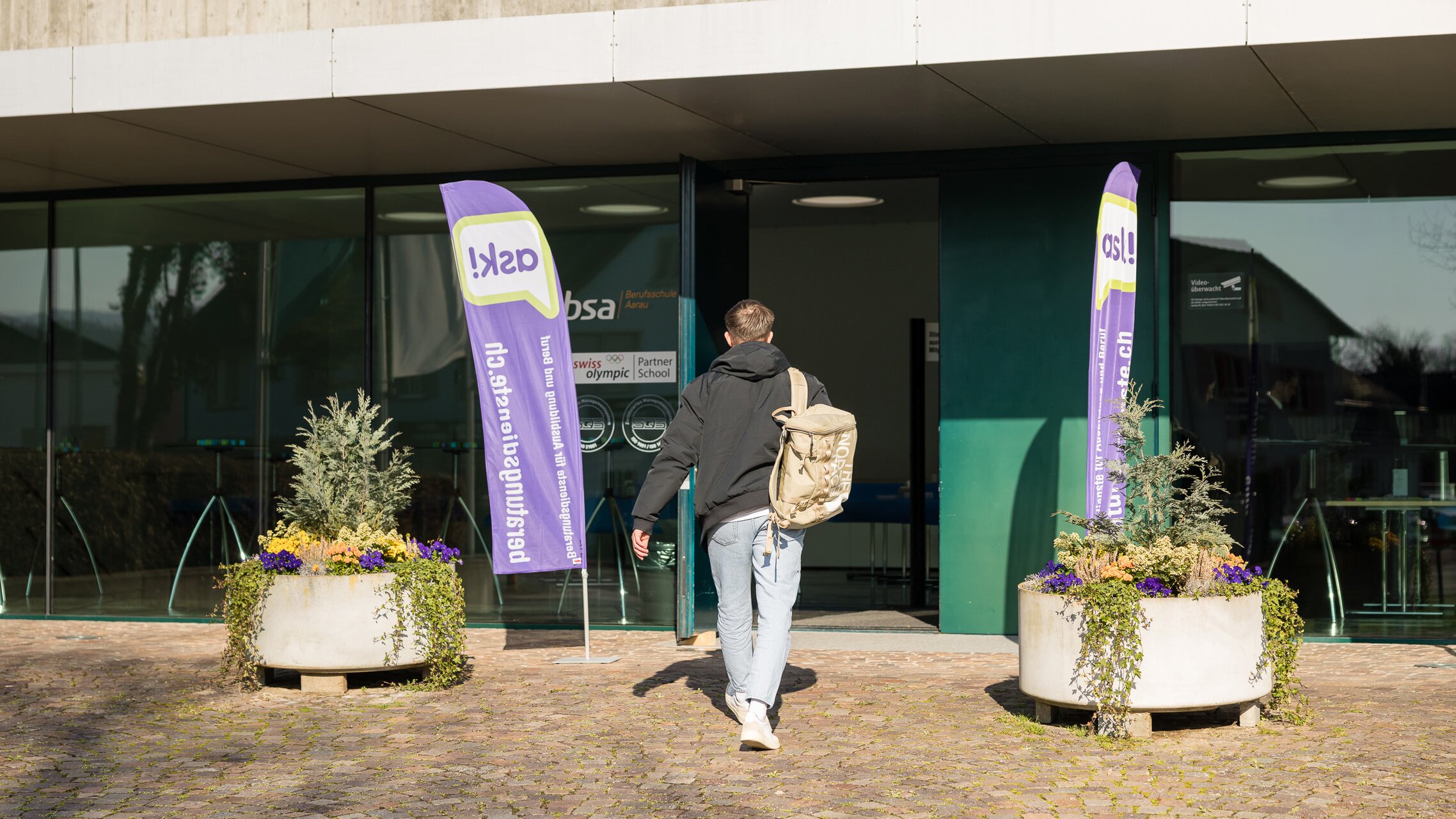 Mann läuft auf den Eingang der Aula zu, der mit ask! Beachflags dekoriert ist und in der die Jubiläumsfeier stattfindet | © ask! / Cornelius Fischer